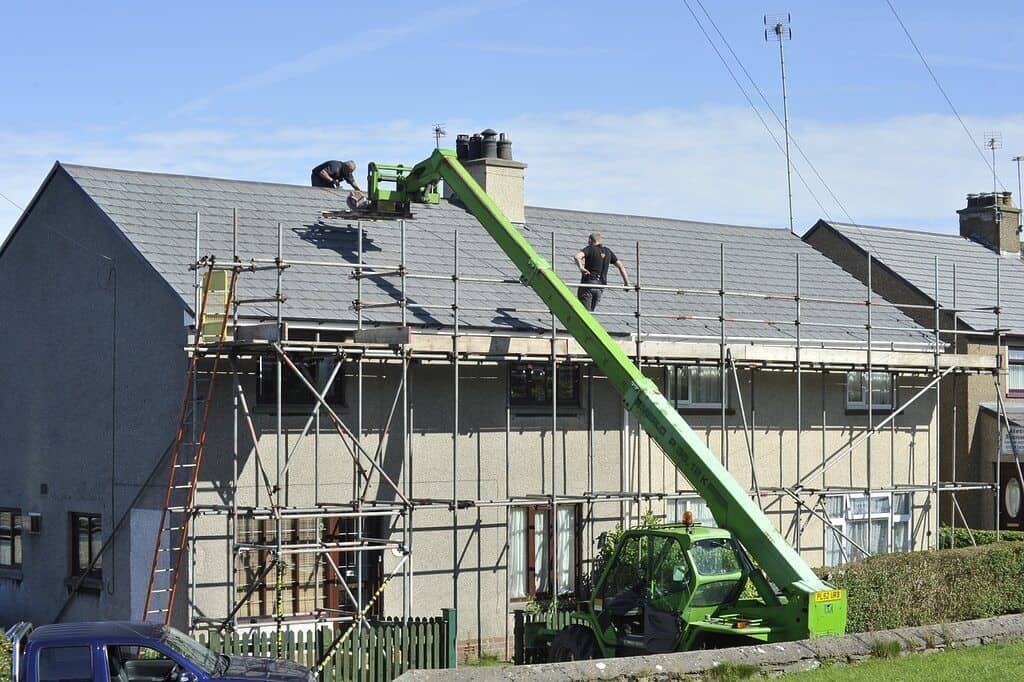 une maison en construction avec un engin vert à côté et deux ouvriers sur le toit