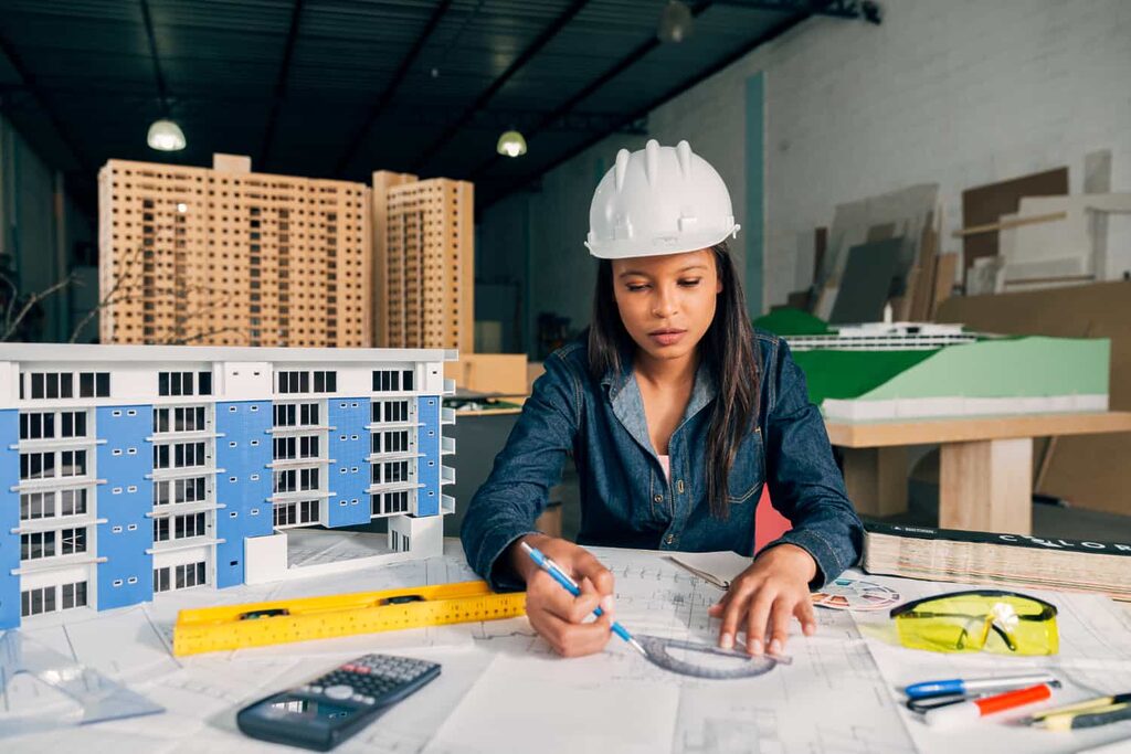 une femme en chemise en jean et un casque de chantier blanc qui travaille sur des plans de construction, avec des maquettes en arrière-plan.