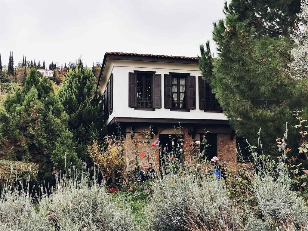 maison traditionnelle à étage blanche avec des volets marrons et un rez-de-chaussée en pierres naturelles, entourée d'arbres et de verdures