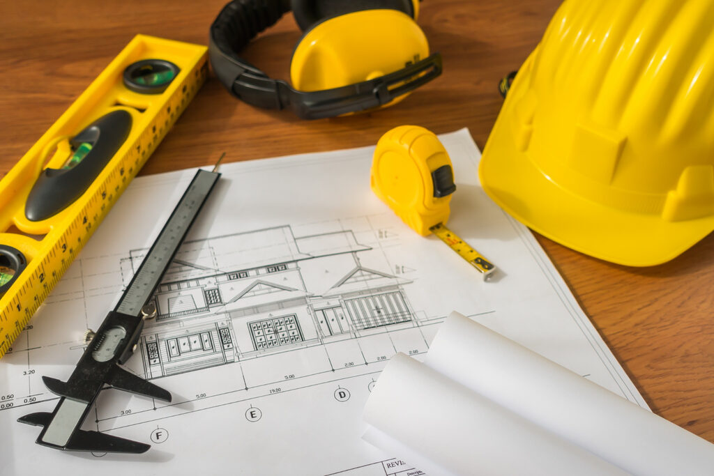 des plans de constructions posés sur une table en bois à côté d'un casque de chantier jaune, un niveau jaune, un mètre jaune, et des oreillettes de chantier. 